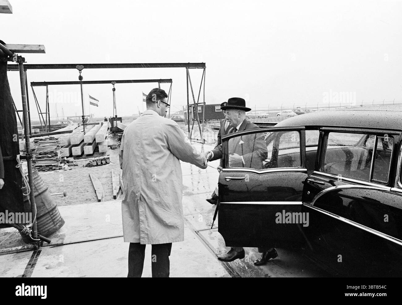 Erster Stapel N.R.P. Tebhin Ned. Raff. Petroleum, Whizgle News, Dutch Desk, Niederlande, 1950 - 2000 am 15-07-1963. Die Abbildung zeigt diese Themen. Die Szene entfaltet sich in einem etwas industriellen Umfeld, das durch gedämpfte Farben und einen bewölkten Himmel gekennzeichnet ist, was auf bewölktes Wetter hindeutet. Links zeigt ein Mann in einem hellen Mantel seine Hand in einer Geste der Begrüßung. Er scheint engagiert und etwas formell zu sein. Ihm gegenüber steht eine weitere Person, die in einem dunklen Anzug und einem legendären schwarzen Hut gekleidet ist und einen klassischen, zeitlosen Stil unterstreicht. Im Vordergrund steht das schlanke, dunkle Fahrzeug Stockfoto