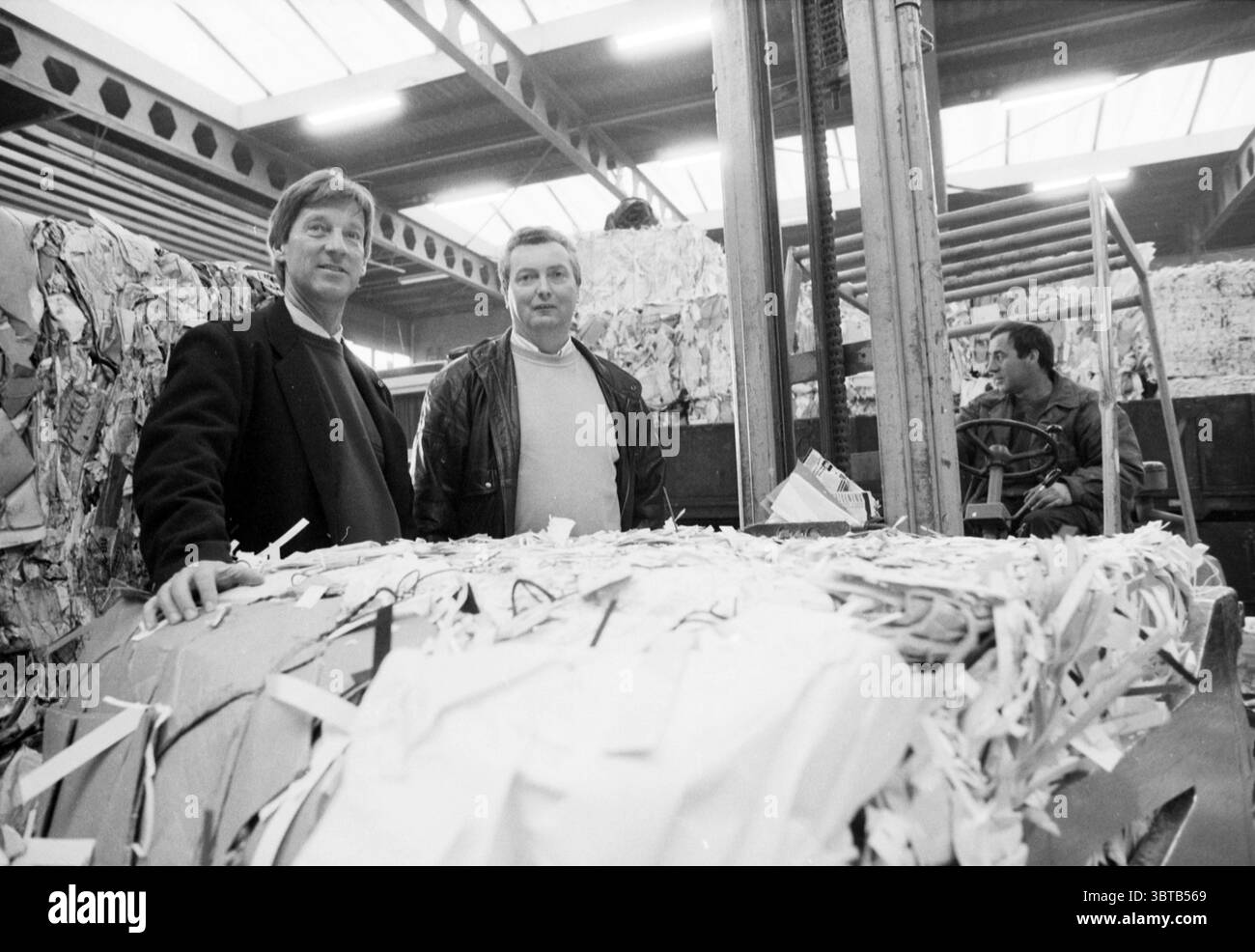 Brantjes und Goebert Brantjes Papier im Zusammenhang mit Jubiläumsjubiläum Jubiläumsjubiläum Paper - Paper Traders etc. 29-03-1989, Whizgle News, Dutch Desk, Niederlande, 1950 - 2000 am 06-04-1989. Dies sind die Themen im Bild. Die Szene zeigt eine geschäftige industrielle Umgebung, die durch große Ballen aus geschreddertem Papier gekennzeichnet ist, die im Vordergrund deutlich gestapelt sind. Zwei Männer stehen eng zusammen und vermitteln ein Gefühl von Kameradschaft in einem Arbeitsumfeld. Der eine Mann trägt eine dunkle Jacke und hat hellbraune Haare und einen nachdenklichen Ausdruck, der andere trägt eine Lederjacke Stockfoto