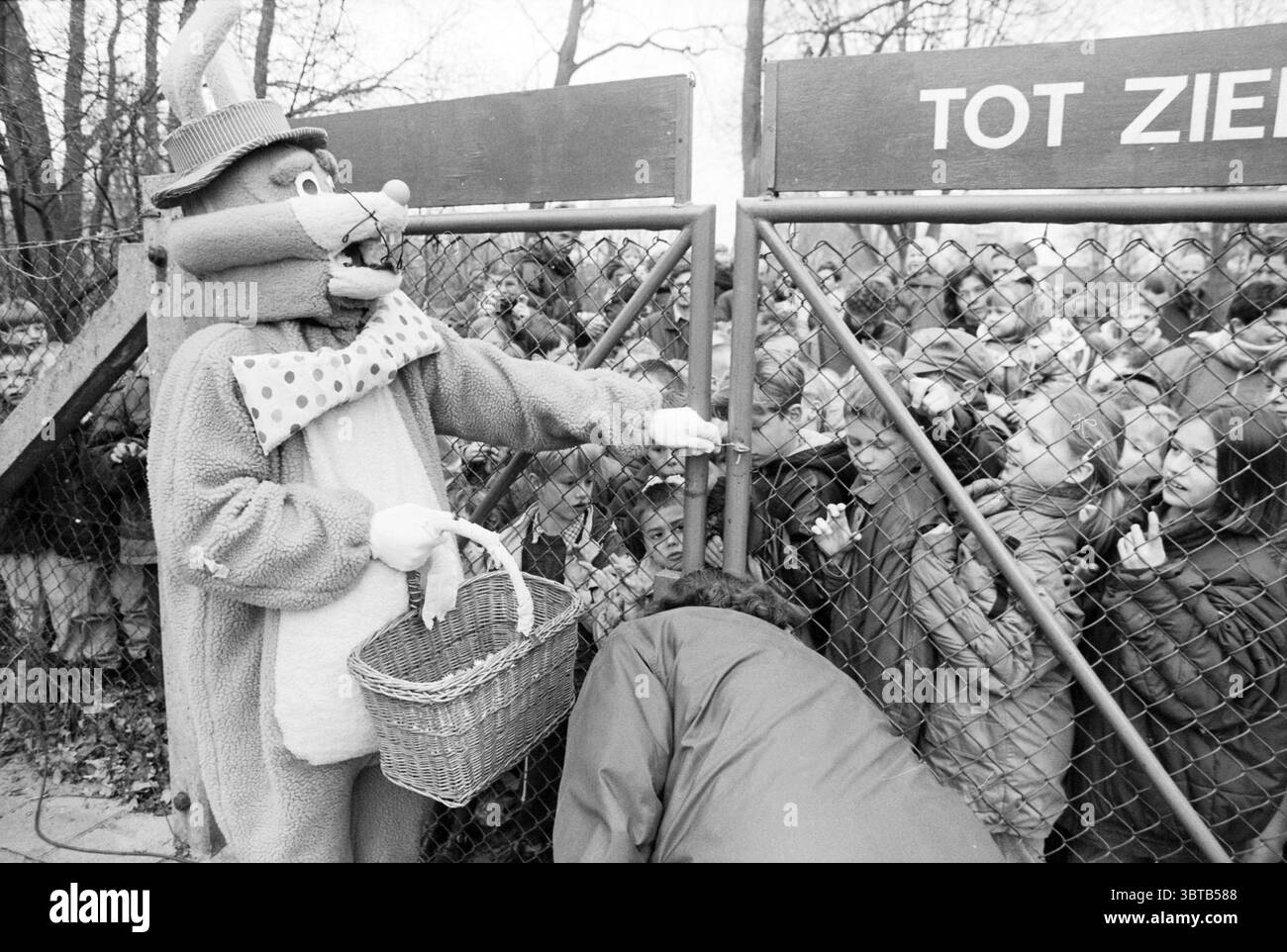 Easter Bunny eröffnet Spielplatz Santpoort Santpoort, Whizgle News, Dutch Desk, Niederlande, 1950 - 2000 am 05-04-1996. Dies sind die Elemente im Bild. In einer lebhaften Umgebung im Freien steht ein skurriler Charakter, der als Kaninchen verkleidet ist, an einem großen Kettenglied. Das Kaninchenkostüm ist flauschig und übertrieben, mit auffälligen Ohren und einem fröhlichen Ausdruck, der sich spielerisch mit der Umgebung abhebt. Die Figur hält einen Korb, der möglicherweise mit Leckereien gefüllt ist, was auf einen festlichen Anlass hindeutet. Hinter dem Tor drückt eine Menge Kinder eifrig gegen den Zaun, ihre Gesichter füllten sich Stockfoto