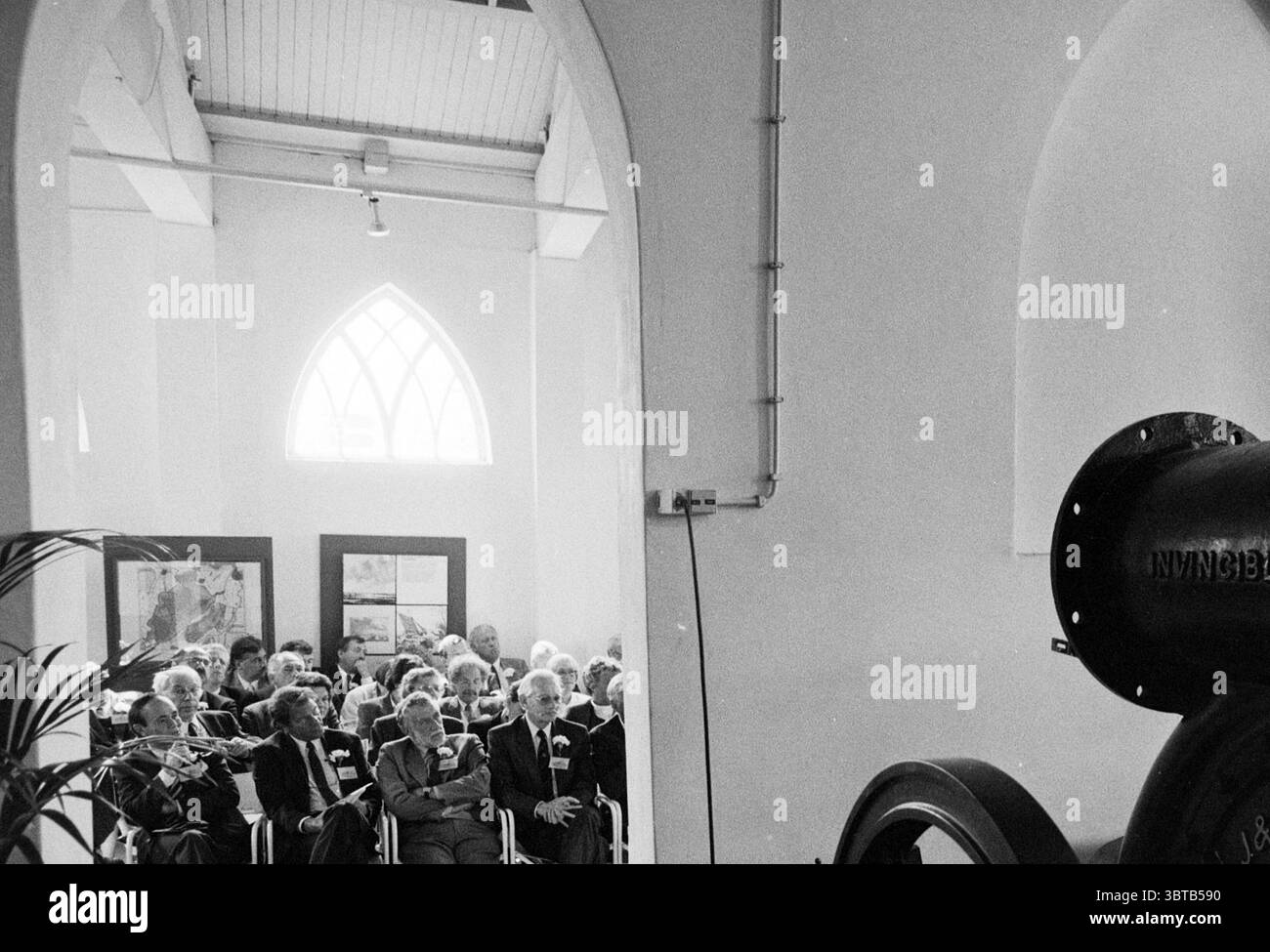 Plakette Cruquius Cruquius, Whizgle News, Dutch Desk, Niederlande, 1950 - 2000 am 18-06-1991. Diese Themen werden in der Abbildung angezeigt. Die Szene entfaltet sich in einem geräumigen, hellen Raum, der sich durch hohe, bogenförmige Decken und große Fenster auszeichnet, die natürliches Licht eindringen lassen und weiche Schatten über den Boden werfen. Die Wände sind hell gestrichen, was die luftige Atmosphäre des Raumes unterstreicht. In Reihen sitzen eine Reihe älterer Menschen, deren Gesichter eine Mischung aus Ausdrücken aufweisen, die auf Engagement und Aufmerksamkeit hinweisen. Sie sind in einer Vielzahl von formellen Kleidungsstücken gekleidet, überwiegend in Farben Stockfoto