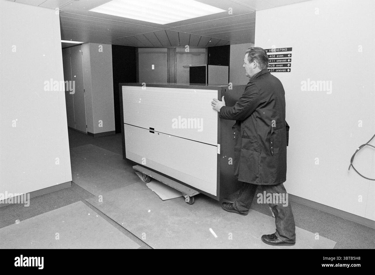 Umzugsbüro Velsen N.A.M. Velsen, Whizgle News, Dutch Desk, Niederlande, 1950 - 2000 am 18.11.1985. Das Bild enthält diese Themen. Die Szene zeigt einen Mann, der ein großes, rechteckiges Objekt auf einem Rollwagen durch einen Flur schiebt. Der Mann trägt einen langen, dunklen Mantel, und sein Ausdruck scheint konzentriert zu sein, während er den schweren Gegenstand manövriert. Der Korridor selbst ist schmal und verfügt über eine neutrale Farbpalette mit weißen Wänden, die zu einer sauberen, fast klinischen Atmosphäre beitragen. Deckenleuchten sorgen für eine helle, gleichmäßige Ausleuchtung, die das Raumgefühl trotz der L Stockfoto