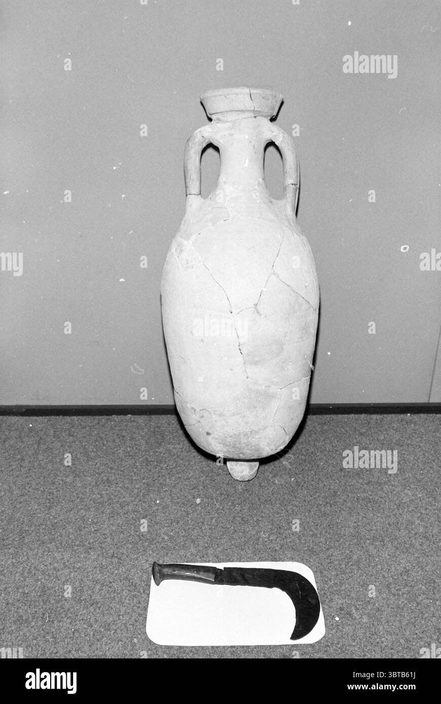 Neue Funde im Kennemer Antiquity Room Amphora + Sichelantiquities, Whizgle News, Dutch Desk, Niederlande, 1950 - 2000 am 01-07-1980. Dies sind die Themen im Bild. In der Szene steht eine große, antike Keramikamphore vor einem schlichten Hintergrund, der ihre historische Bedeutung unterstreicht. Die Amphore hat einen glatten, abgerundeten Körper mit leichten Einbuchtungen in der Nähe der Basis und zeichnet sich durch einen stilisierten Hals aus, der in zwei breite Griffe mündet und ihr ein elegantes Profil verleiht. Die Oberfläche der Amphore erscheint verwittert und zeigt eine Palette gedämpfter Erdtöne – variierend Stockfoto