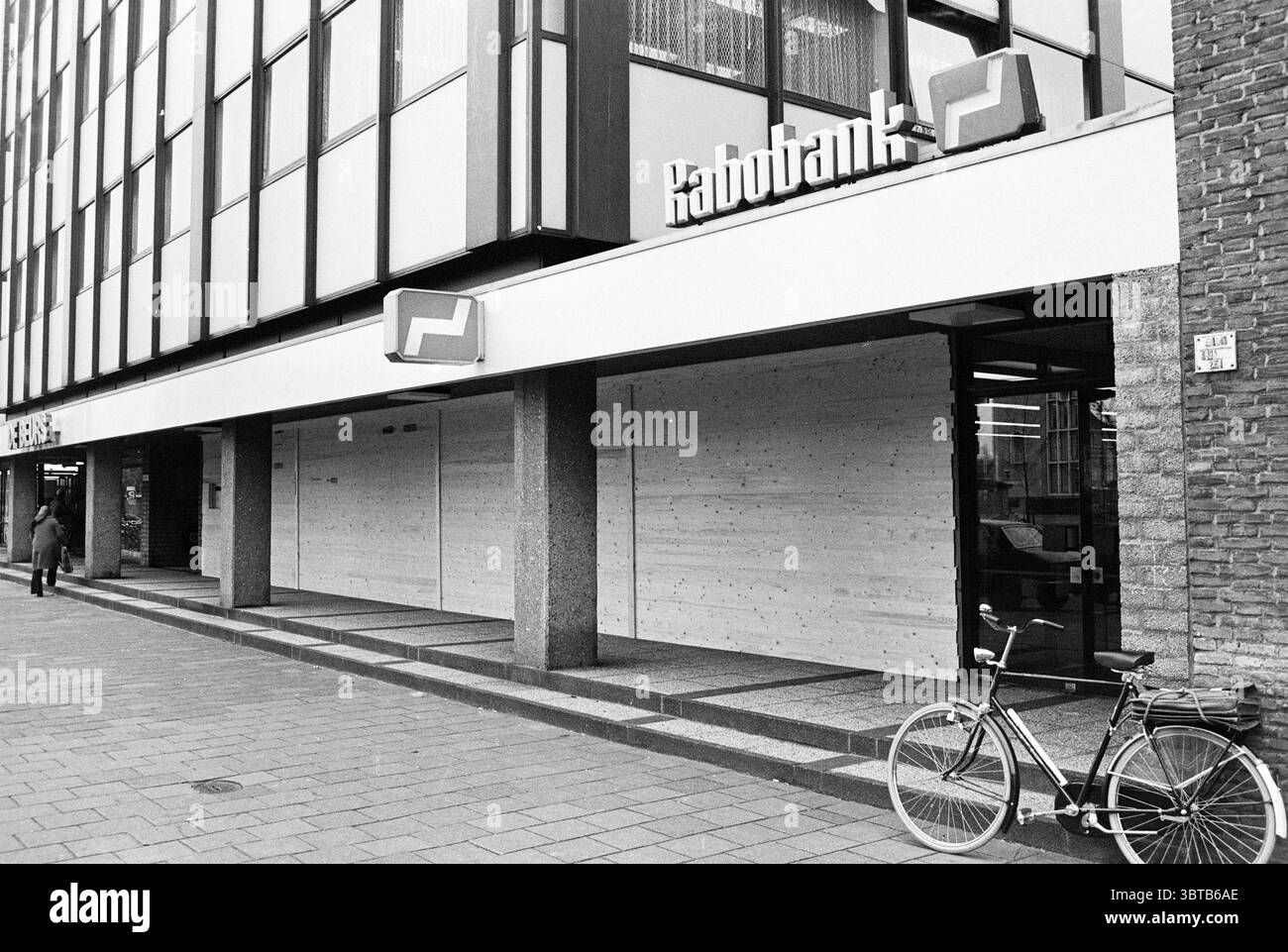 Fenster mit Glasverkleidung, Rabobank IJmuiden. Buntglas bläst Glas im Allgemeinen, Whizgle News, Dutch Desk, Niederlande, 1950 - 2000 am 03-03-1977. Diese Themen werden in der Abbildung angezeigt. Die Szene zeigt ein modernes Gebäude, das für sein schlankes und minimalistisches Architekturdesign bekannt ist. Die Fassade verfügt über eine Reihe von großen, reflektierenden Fenstern und einen breiten Eingang, der ihr eine offene Atmosphäre verleiht. Die Farbpalette wird von Grau-, weiß- und Schwarztönen dominiert, was die schlichte, urbane Atmosphäre unterstreicht. Im Vordergrund steht ein klassisches Fahrrad mit einem einfachen Rahmen und einem Gang am Gebäude Stockfoto