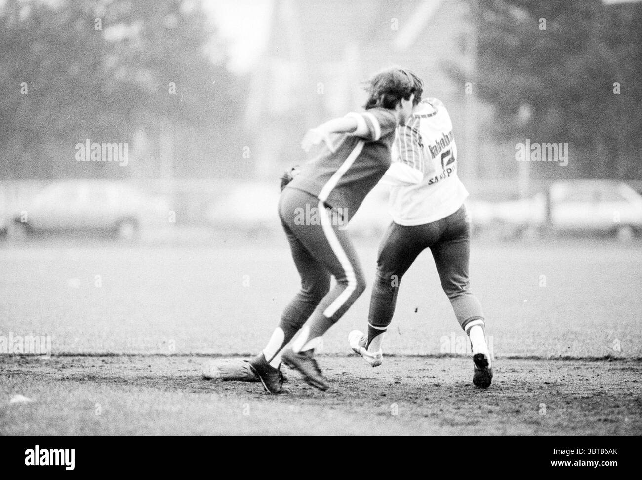 DSS - Terrasvogels Softball, Whizgle News, Dutch Desk, Niederlande, 1950 - 2000 am 28-08-1980. Diese Themen sind in der Abbildung dargestellt. In einer lebendigen Sportszene sind zwei Spieler in einem dynamischen Moment aktiv und demonstrieren Bewegung und Interaktion. Ein Athlet trägt eine rote Uniform, die von weißen Streifen an den Seiten gekennzeichnet ist, während der andere mit einem weißen Oberteil mit einem Markenlogo und einer dunkleren Hose gekleidet ist. Die Umgebung scheint ein grasbewachsenes Feld zu sein, möglicherweise ein Sportplatz, mit leicht erhöhten Bereichen, die auf eine ländliche Kulisse hinweisen. Die Atmosphäre ist etwas bewölkt, Stockfoto