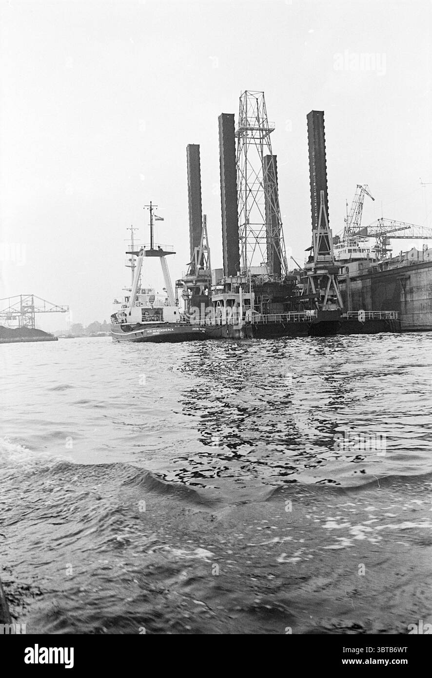 Vorbereitung der Abreise Jacob van Heemskerk Wijsmuller, Whizgle News, Dutch Desk, Niederlande, 1950 - 2000 unter 30-10-1968. Das Bild enthält diese Themen. Die Szene fängt ein geschäftiges maritimes Industriegebiet ein, das durch eine Mischung aus großen Strukturen und Schiffen gekennzeichnet ist. Im Vordergrund wird ein Binnenschiff oder Boot angedockt und zeigt einen robusten Rumpf, der in weiß- und Dunkelblau lackiert ist, was eher auf funktionellen Nutzen als auf ästhetische Details hindeutet. Das Schiff ist mit hoch aufragenden Metallkranen oder Bohranlagen ausgestattet, die majestätisch ansteigen, wobei ihre starken vertikalen Linien sich von der weichen Linie abheben Stockfoto