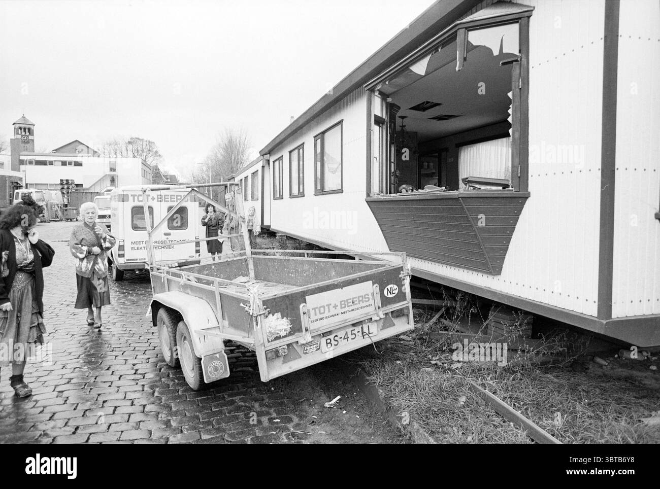 Unfallkarawane Velsen-Noord Velsen-Noord, Whizgle News, Dutch Desk, Niederlande, 1950 - 2000 am 29-11-1990. Das Bild enthält diese Themen. Die Szene zeigt eine desolate Außenumgebung neben einem teilweise beschädigten Gebäude. Das Gebäude, das ein großes Fenster und eine unregelmäßig geformte Öffnung aufweist, scheint eine bewegliche Struktur oder ein provisorisches Wohnhaus zu sein. Die Wände sind in gedämpften Tönen gemalt, mit einem einfachen Design, das andeutet, dass es einst ein lebhafteres Aussehen hatte. Im Vordergrund sitzt ein Metallanhänger schräg gegen den unebenen Boden, dessen Oberfläche leicht rostet und gelagert ist Stockfoto