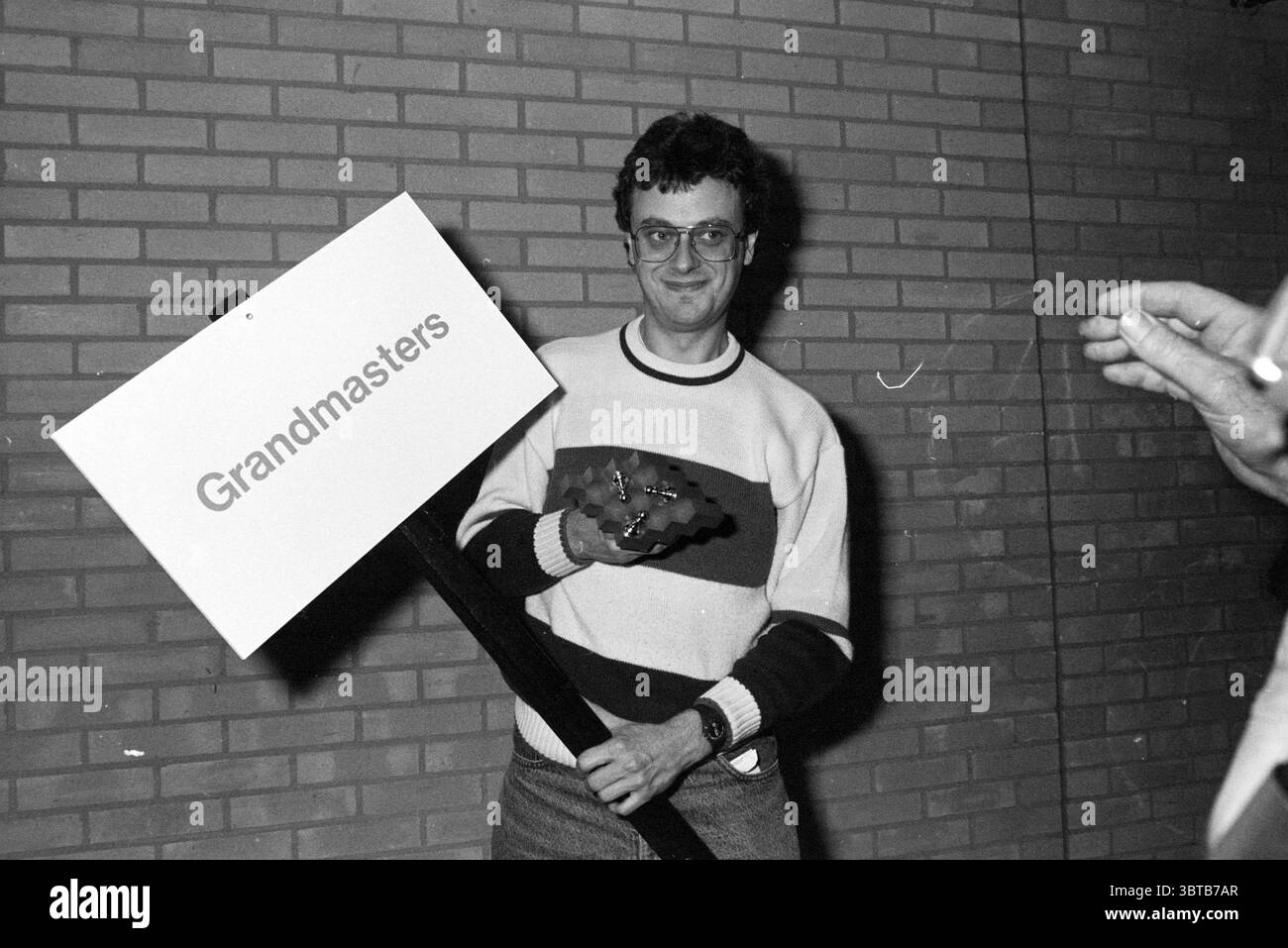 Verpflegung und Preisverteilung H.S.T. HST, Whizgle News, Dutch Desk, Niederlande, 1950 - 2000 am 28-01-1990. Diese Themen sind in der Abbildung dargestellt. Im Vordergrund steht eine Person mit einem großen Schild mit einer einfachen Botschaft, das zur besseren Sichtbarkeit zentral positioniert ist. Die Person, die in einem horizontal gestreiften Pullover mit kräftigen Farben weiß, Rot und Blau gekleidet ist, scheint ein verspieltes Auftreten zu haben, ergänzt durch ein leichtes Lächeln. Sie haben eine Brille und ein allgemeines Aussehen des Genusses. Ihre andere Hand hält ein Objekt, das einer kleinen Trophäe oder Auszeichnung ähnelt und ein Element der Leistung hinzufügt Stockfoto