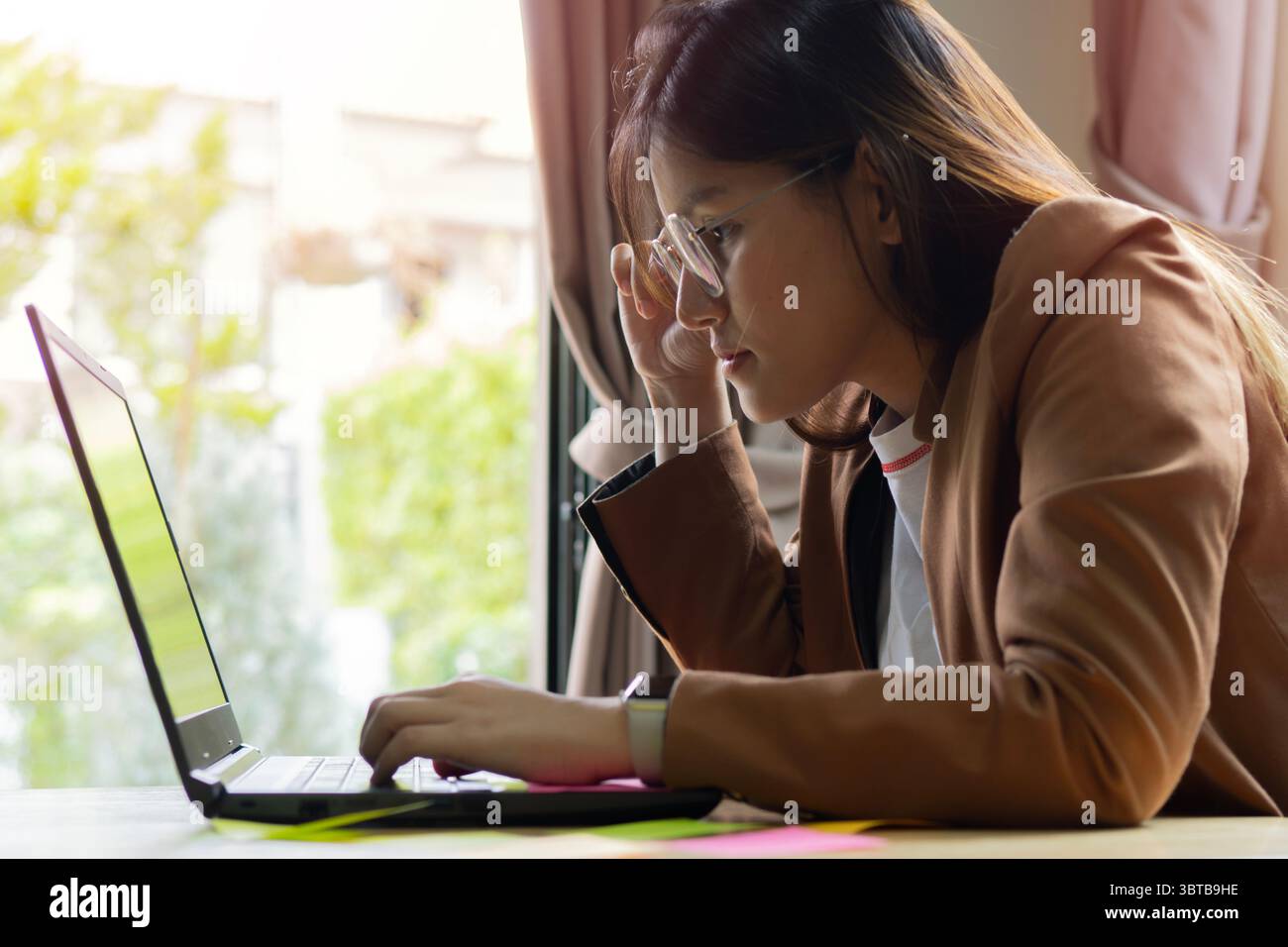 Junge, schöne asiatische Frau mit Brille, die im Büro am Computer arbeitet. Geschäftskonzept. Seitenansicht. Stockfoto