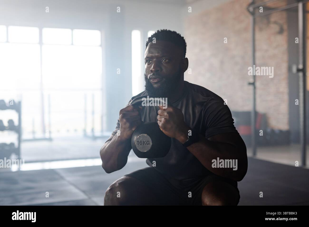 Afroamerikaner, der 16 kg Kettlebell hält, hockt auf Gummiboden im Fitnessstudio mit Hantelständer Stockfoto