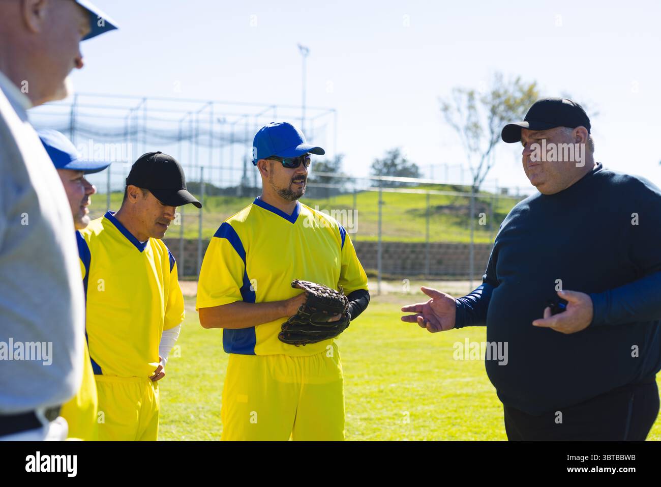 Das Baseballteam der Männer hält Handschuhe in gelben Uniformen und hört dem Trainer auf dem Baseballfeld zu Stockfoto