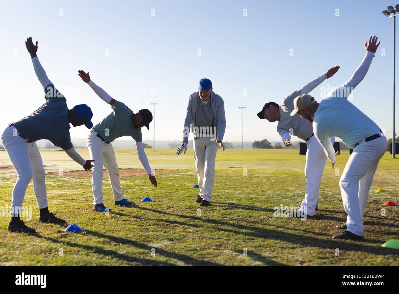 Verschiedene männliche Mannschaftskameraden dehnen sich seitlich auf dem Grasfeld und tragen Baseballkappen in der Nähe von Trainingskegeln Stockfoto