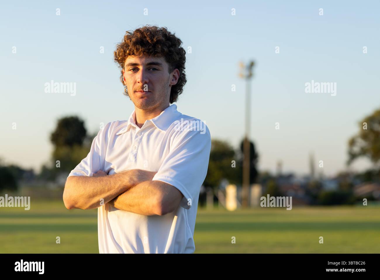 Männlicher Athlet, der die Arme auf dem Sportplatz unter Flutlicht in goldener Stunde kreuzt, trägt ein weißes Poloshirt Stockfoto