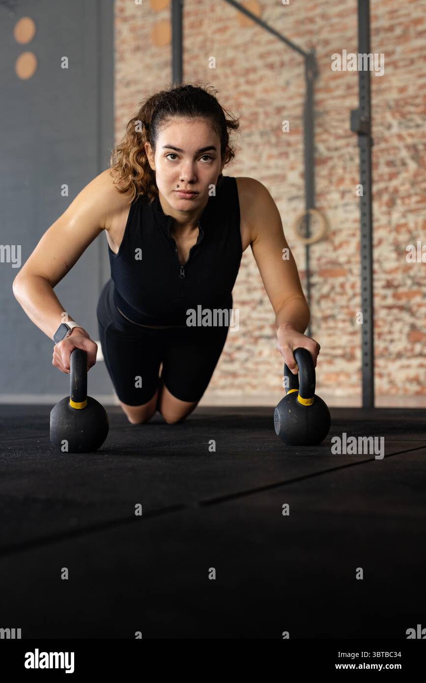 Weibliche Athletin mit Liegestützvariante auf Kettlebells im Fitnessstudio mit Fitnessband am Handgelenk Stockfoto