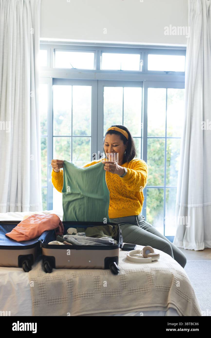 Asiatische Frau in den späten 30er Jahren auf dem Bett im Schlafzimmer, die Koffer packte und die grüne Oberseite neben Kopfhörern untersucht Stockfoto