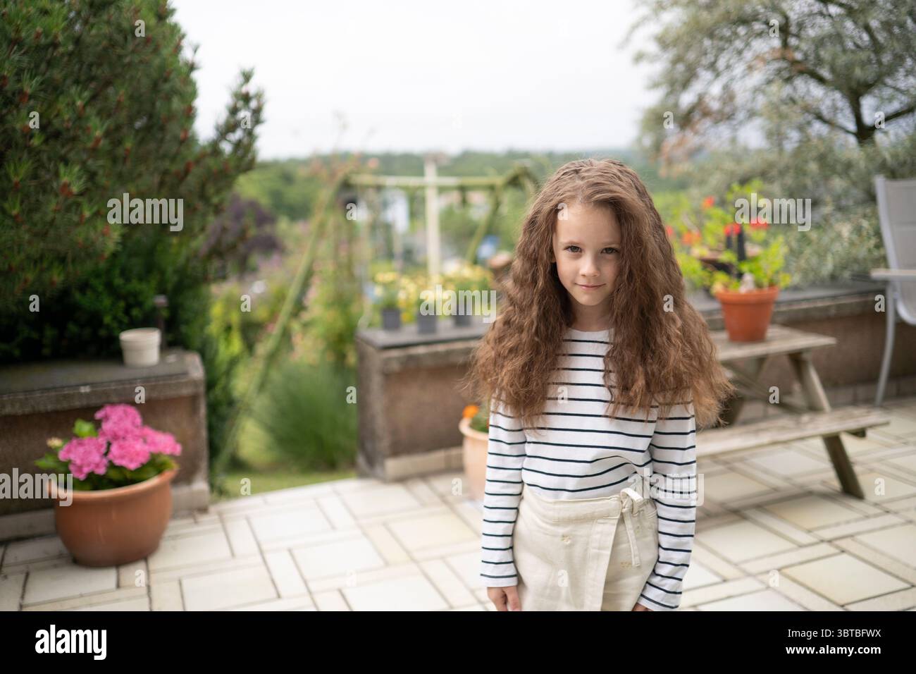 Lächelndes Mädchen in gestreiftem T-Shirt auf einer grünen Sommerterrasse. Konzept: Sorgenfreier Geist, warme Tage, Gartenleben, verspielte Energie. Stockfoto
