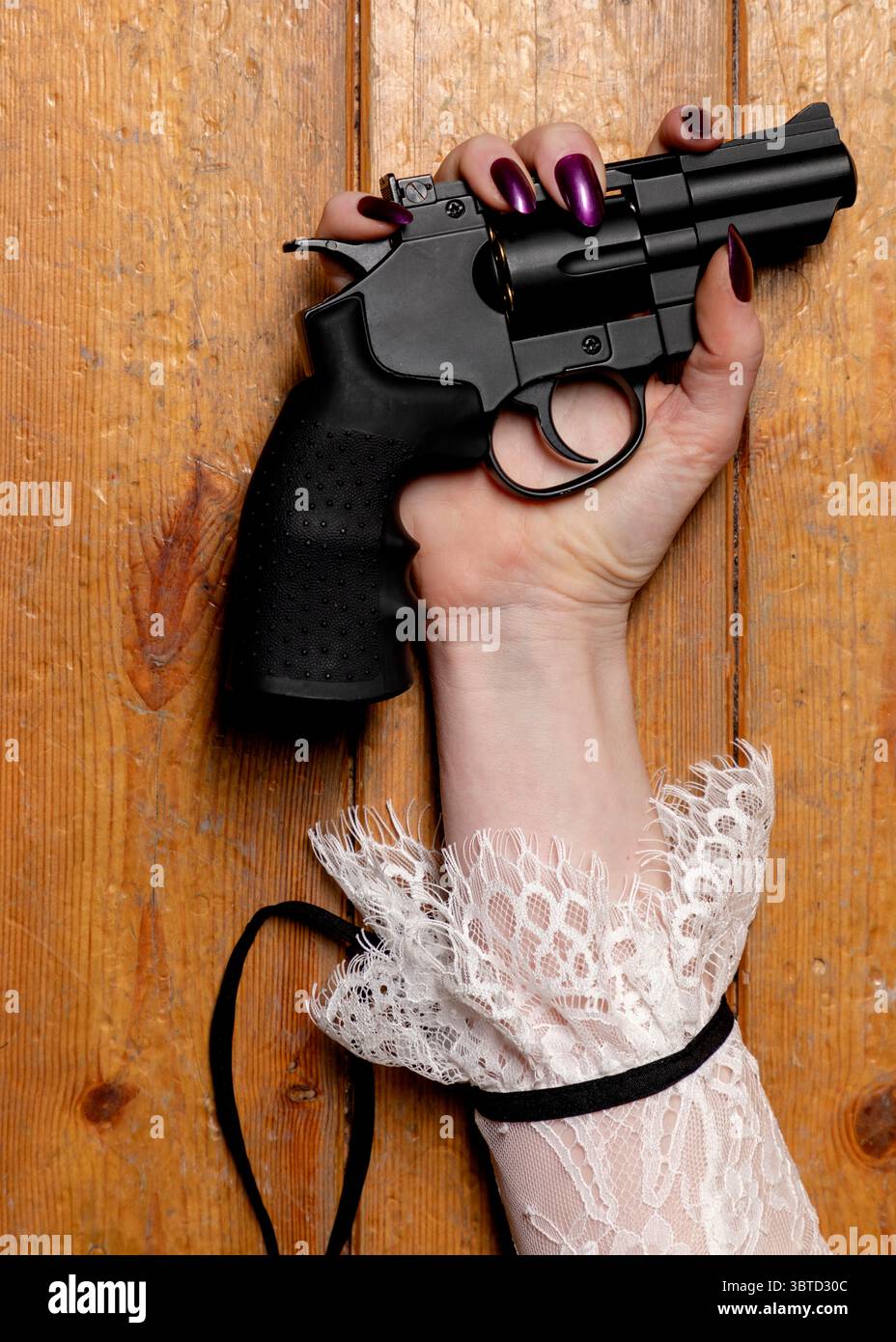 Eine Hand mit violettem nagellack hält einen schwarzen Revolver vor einem hölzernen Hintergrund. Das Handgelenk ist mit einer weißen Spitzenmanschette und einem schwarzen Band verziert. Stockfoto