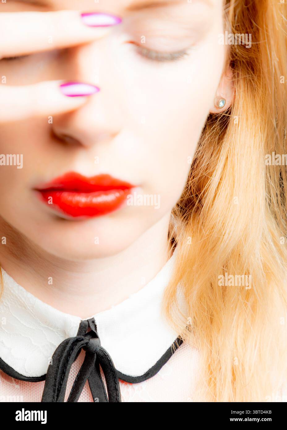 Eine Nahaufnahme einer Frau mit roten Lippen, geschlossenen Augen und einer Hand, die ihr Gesicht bedeckt. Sie hat lange blonde Haare und trägt ein weißes Hemd mit Kragen Stockfoto