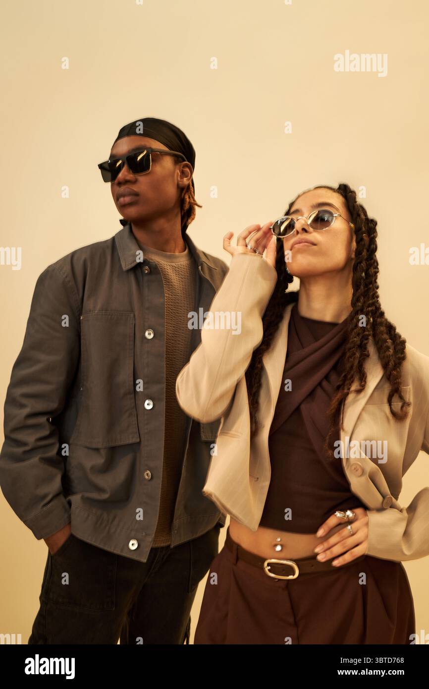 Junger Schwarzer Mann und junge Schwarze Frau, die eine Sonnenbrille tragen, posieren selbstbewusst vor einem schlichten Hintergrund, Frau, die die Sonnenbrille anpasst, während sie beide von der Kamera wegschauen, stilvolle Einstellung Stockfoto