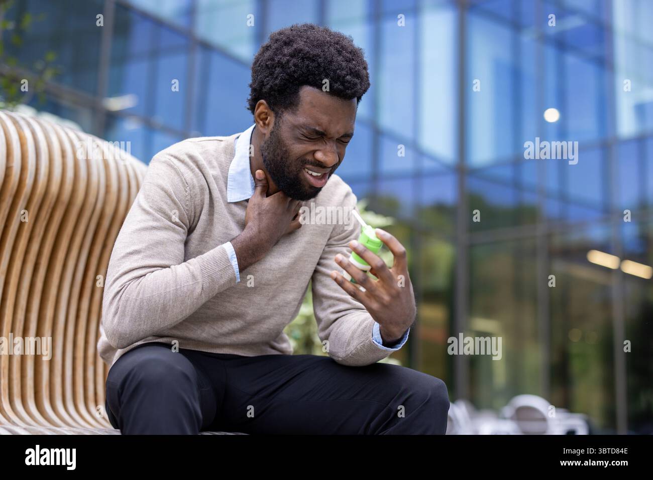 Ein afroamerikanischer Mann leidet unter Halsschmerzen und benutzt draußen ein Kehlspray, in der Nähe eines modernen Glasgebäudes. Stockfoto