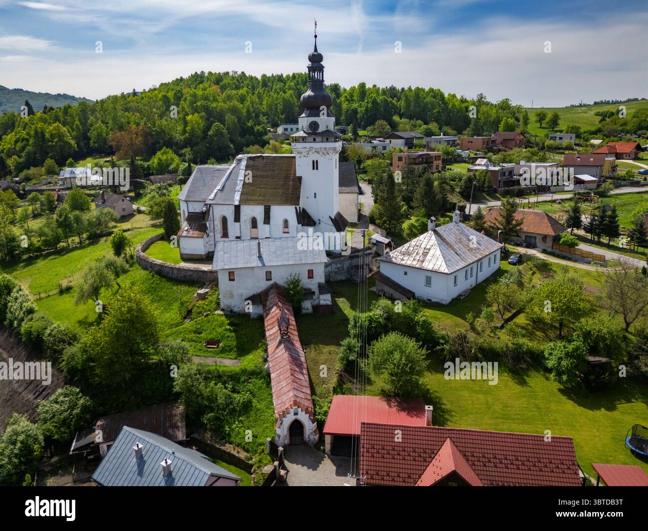 Blick aus der Vogelperspektive auf die weiße Kirche mit dunklem Dach, eine Brücke und umliegendes Grün, das mit den fernen Hügeln verschmilzt, Banskobystrický, Banská Belá kraj, Slowakei. Stockfoto