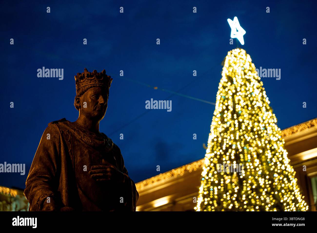 Eine Bronzestatue steht in Pamplona, Navarra, España, mit einem wunderschön beleuchteten Weihnachtsbaum, der hell vor dem tiefen Abendhimmel leuchtet. Das fest Stockfoto