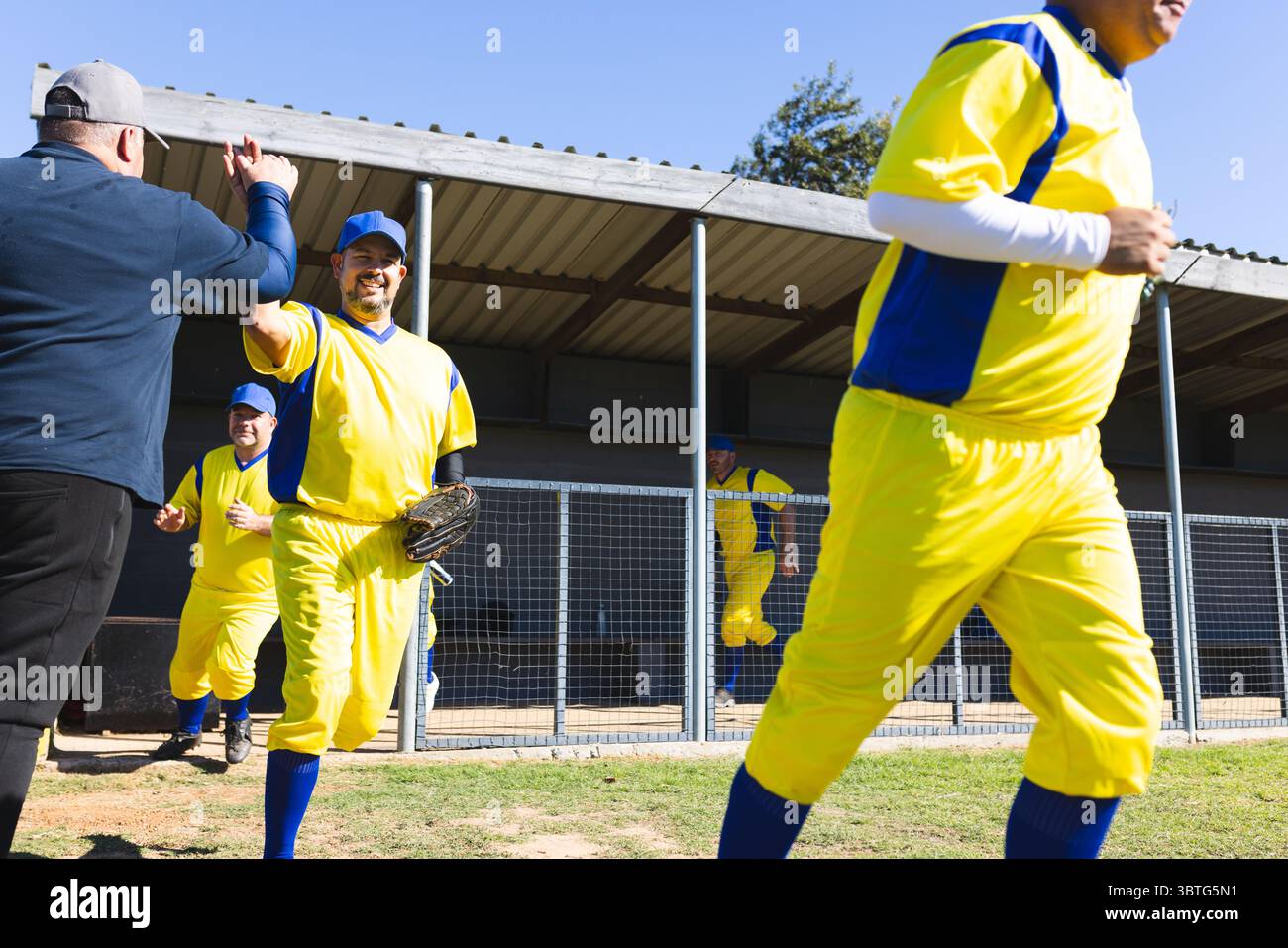 Verschiedene männliche Baseballspieler in gelben Trikots High-Fiving-Trainer, während sie Handschuhe in der Nähe des Dugouts halten Stockfoto