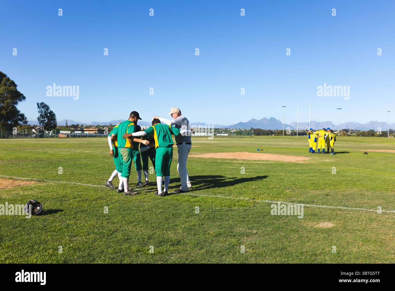 Männliche Baseballspieler und Coach, die sich auf dem Baseballfeld mit dem Helm von Catcher auf Pitcher's Mound tummeln Stockfoto