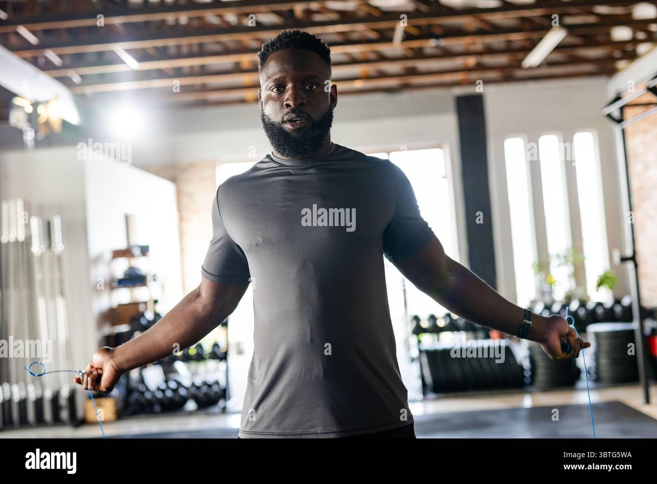 Afroamerikaner, der Trainingsausrüstung trägt und im Fitnessstudio unter hohen Fenstern mit Gewichtshaltern ein Seil überspringt Stockfoto