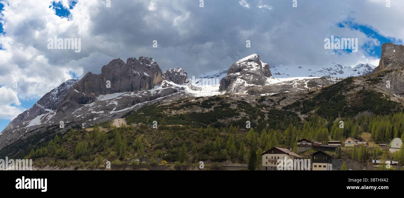 Panoramablick auf die majestätischen schneebedeckten Gipfel der Marmolada, die sich über einer üppigen grünen Landschaft mit verstreuten Alpenhäusern in den Dolomiten erheben. Stockfoto