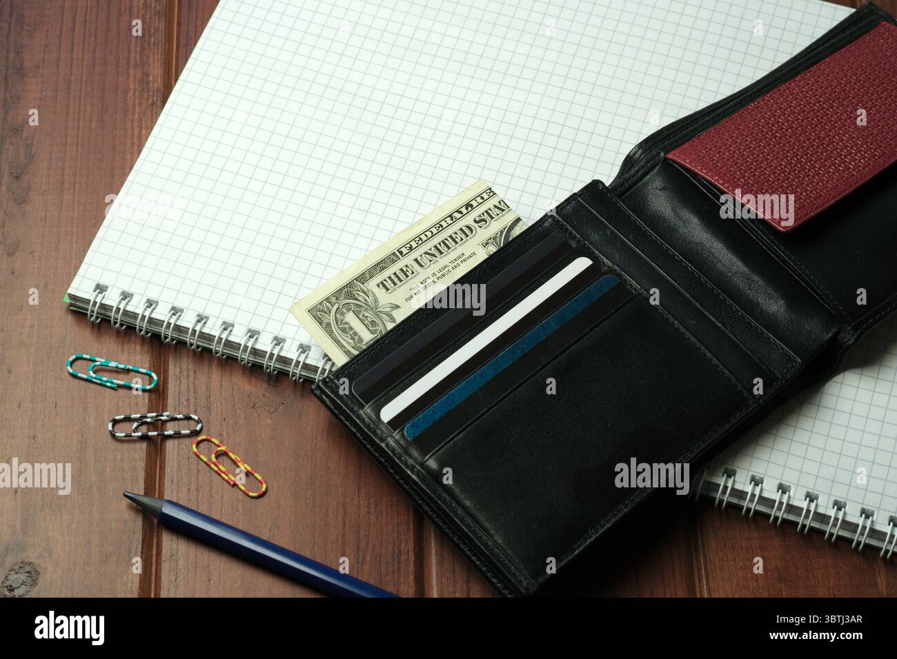 Eine schwarze Geldbörse zeigt Bargeld und Karten neben Spiralnotizbüchern und farbenfrohen Büroklammern. Stockfoto
