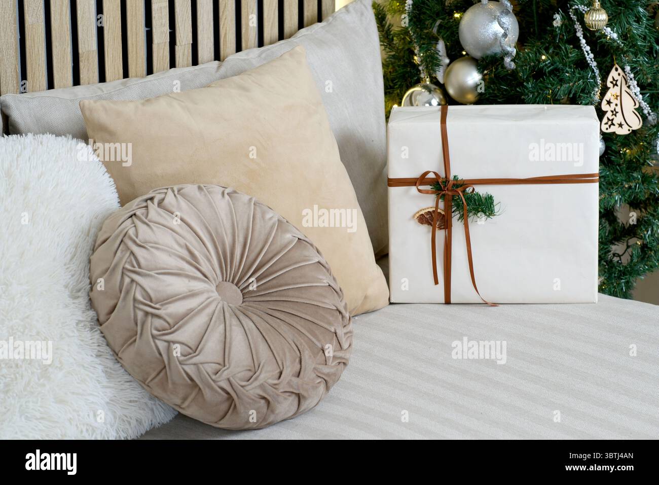 Weiche Kissen und ein wunderschön gewickeltes Geschenk neben einem festlichen Baum sorgen für Wärme. Stockfoto