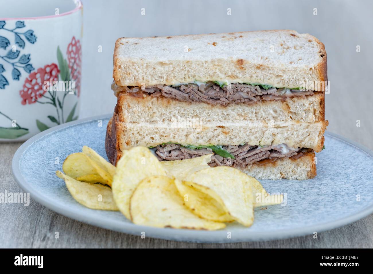 UK. Ein großzügiges Rindersandwich, serviert auf Weißbrot mit Salat und Mayo-Dressing. Es wird auf einem Teller mit Chips und einem heißen Getränk serviert Stockfoto