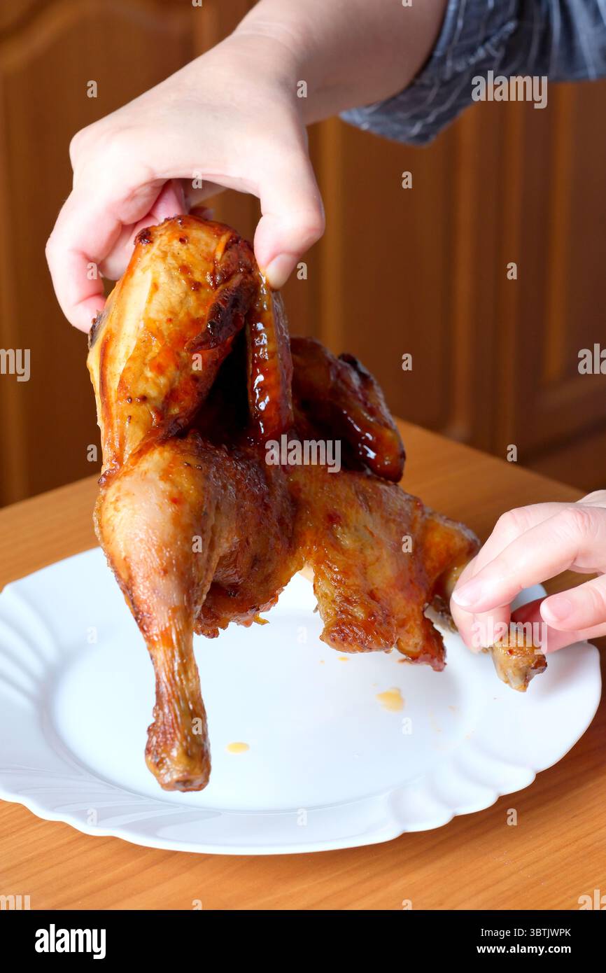 Die Hände einer älteren Frau bedienen ein gegrilltes Huhn und zeigen es in der Küche an einem braunen Tisch, ohne Gesicht, Nahaufnahme Stockfoto