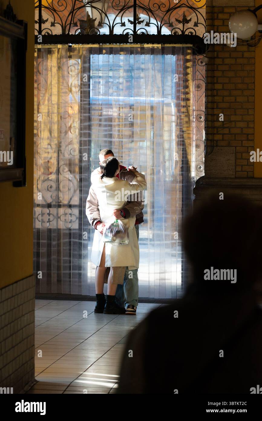 [Budapest], [Ungarn] - 07. Oktober 2025: Blick auf eine intime Umarmung, eingerahmt von kunstvollen Eisenarbeiten und knalligen Vorhängen, die die Szene in einem klassischen Budapester Gebäude mit sanftem Licht beleuchten. Stockfoto