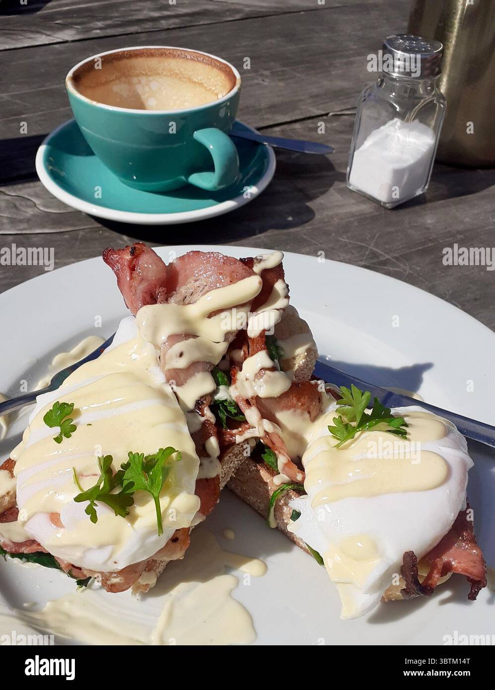 Eggs Benedict mit knusprigem Speck, pochierten Eiern und Hollandaise-Sauce serviert auf Toast und Kaffee auf einem Holztisch in natürlichem Licht Stockfoto