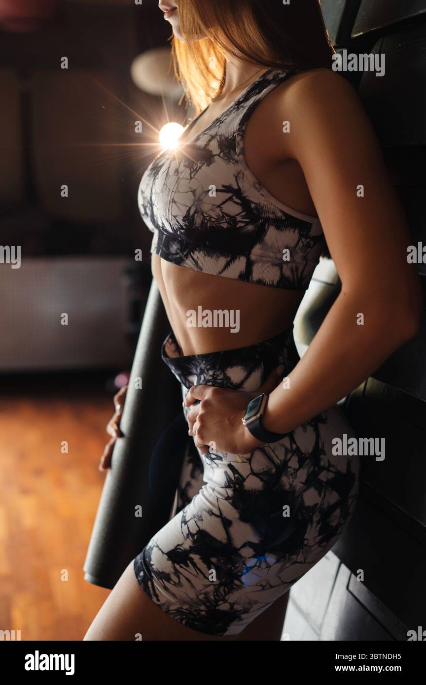 Eine Frau in einem stilvollen Workout-Outfit hält eine Yogamatte in einem schwach beleuchteten Raum mit Holzboden. Das Sonnenlicht erzeugt ein warmes Leuchten, Highlight Stockfoto