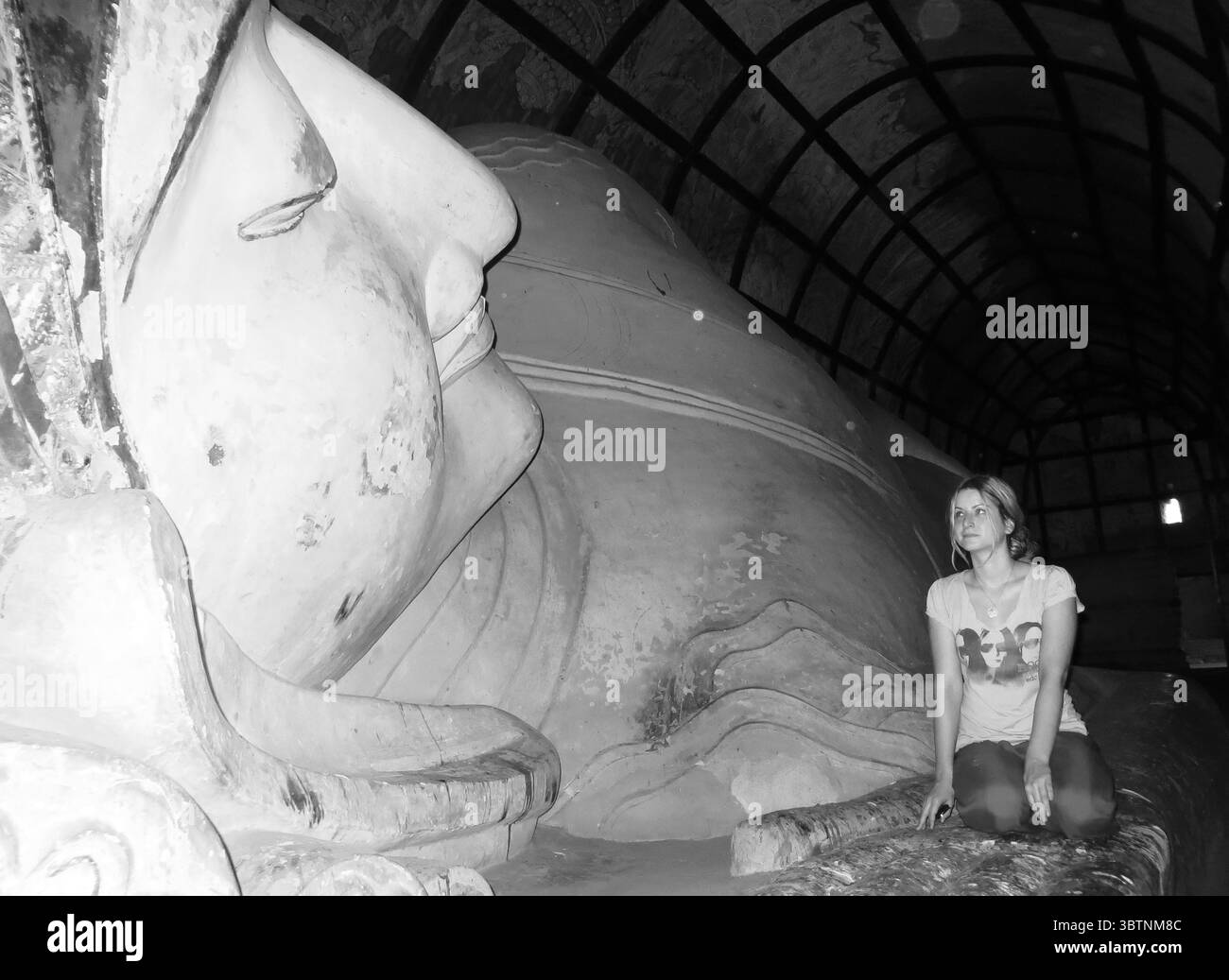 Schwarz-weiß-Foto einer jungen Frau, die neben der massiven liegenden Buddha-Statue im schwach beleuchteten Tempel Bagan Myanmar kniet Stockfoto