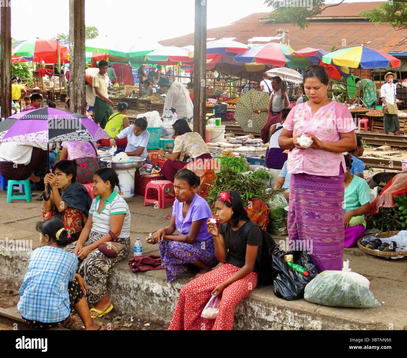 Geschäftiger Markt im Freien an einem Bahnhof in Myanmar mit Händlern und Einkäufern inmitten farbenfroher Stände und traditioneller Kleidung. Stockfoto