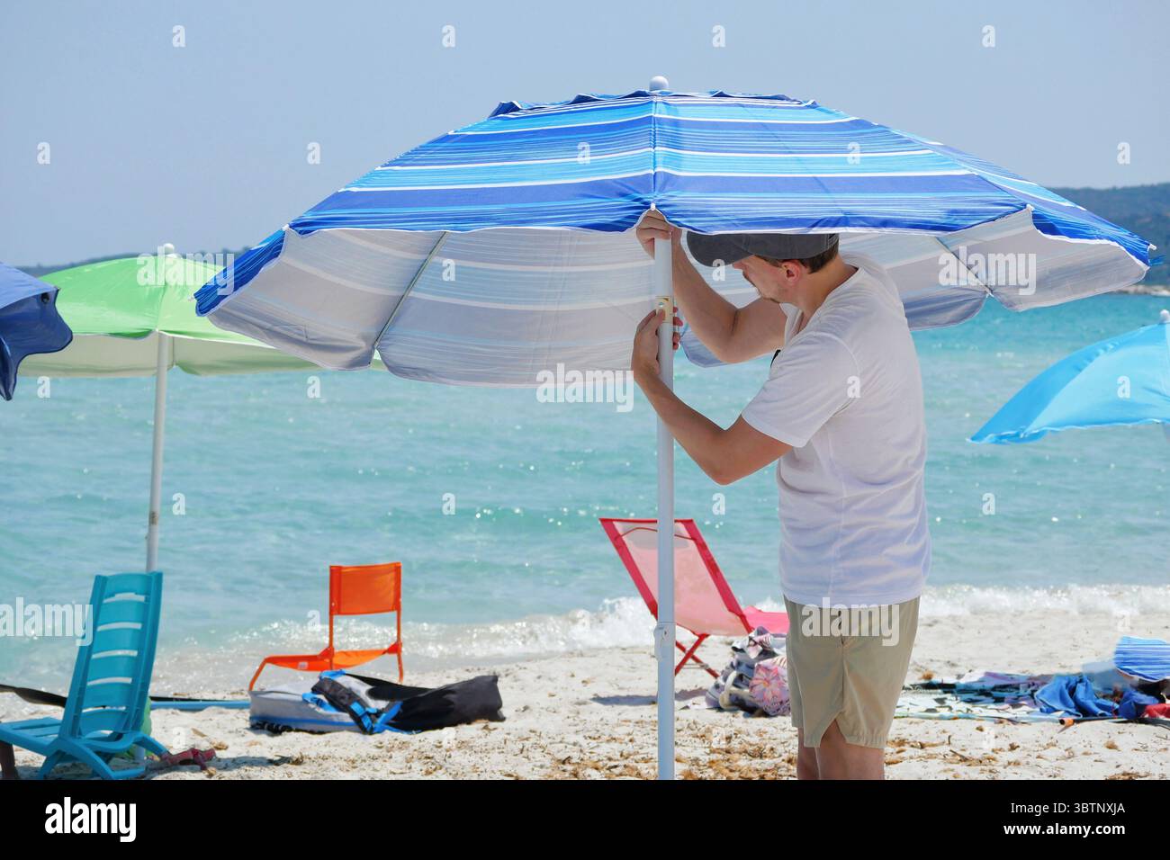 Junger Mann, der an einem sonnigen Sommertag einen Sonnenschirm aufstellt. Reisen, Sommerferien, Outdoor-Freizeitangebote und Beach Lifestyle Konzept. Stockfoto