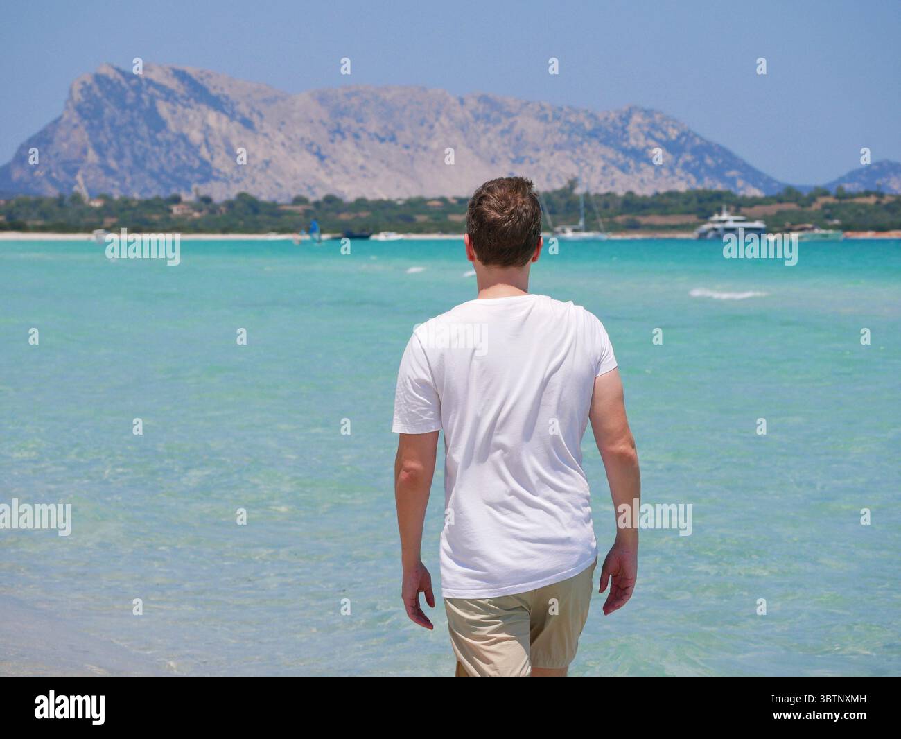 Ein junger Mann in Sonnenbrille und weißem T-Shirt, der am Hintergrund des türkisfarbenen Meeres an einem tropischen Strand steht. Lifestyle, Reisen, Mode und Sommer Ho Stockfoto