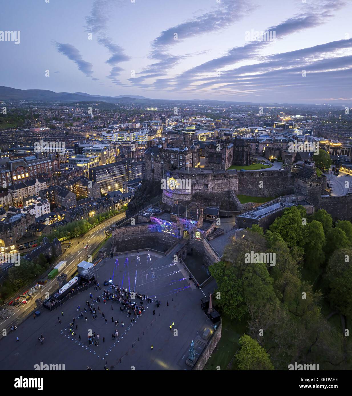 Edinburgh, Vereinigtes Königreich - 16. Juni 2025: Aus der Vogelperspektive das Edinburgh Castle unter dem Dämmerhimmel, seine alten Steinmauern beleuchtet in einer Stadtlandschaft, wo Lichtschichten vor der eindringenden Dämmerung glitzern, während die leuchtenden Grüntöne der umliegenden Bäume einen scharfen Kontrast zum grauen Stein und den weit entfernten Hügeln bilden, die in den Horizont fallen, und Figuren im Vordergrund versammelt sind. Stockfoto
