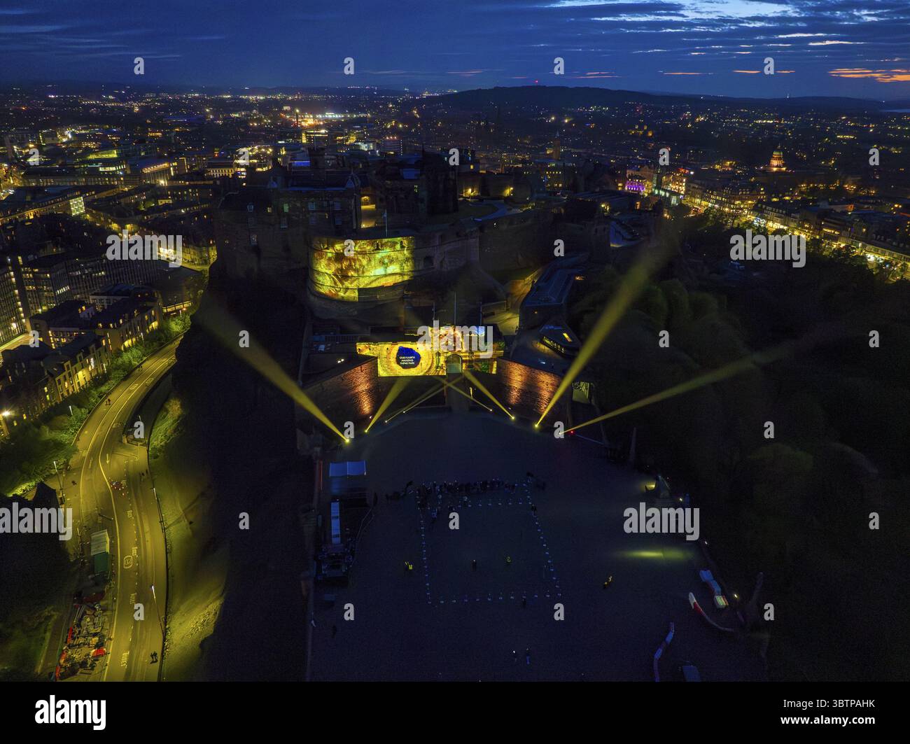 Edinburgh, Vereinigtes Königreich - 16. Juni 2025: Nächtlicher Blick auf Edinburgh Castle, wo leuchtende gelbe Scheinwerfer durch die Dunkelheit schneiden, im Kontrast zu den gedämpften Lichtern der Stadt unter einem Dämmerhimmel. Stockfoto