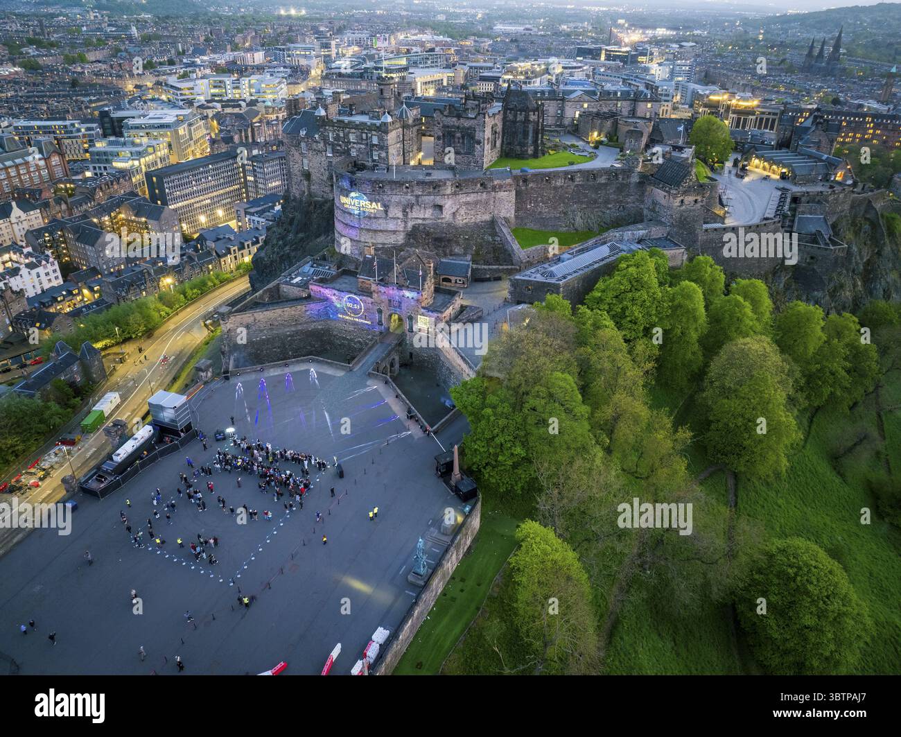 Edinburgh, Vereinigtes Königreich - 16. Juni 2025: Aus der Vogelperspektive auf dem Castle Rock thront das majestätische Edinburgh Castle mit seinen alten Steinmauern, die sanft im schwindenden Licht leuchten, gegenüber dem lebhaften Grün der umliegenden Bäume und dem städtischen Wandteppich der Stadt unten, wo sich winzige Figuren auf der Esplanade versammeln, die von einem Lichterkonstellation beleuchtet werden, wenn die Dämmerung absinkt. Stockfoto