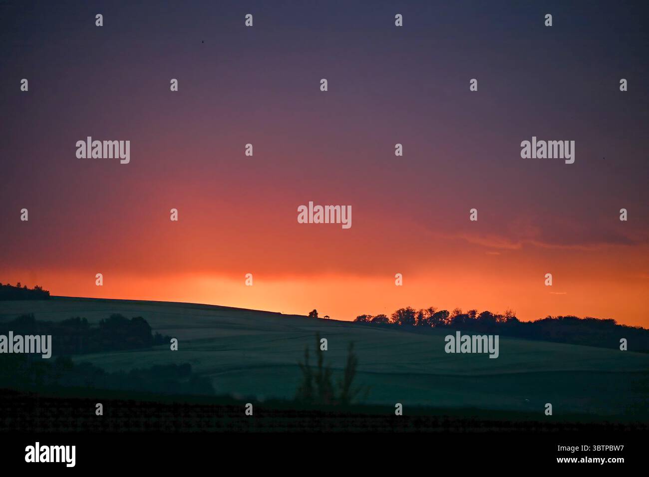 Dunkle, sanfte Hügel mit Bäumen, die sich vor einem leuchtenden roten und orangen Sonnenuntergangshimmel in tiefviolette Töne verwandeln. Stockfoto