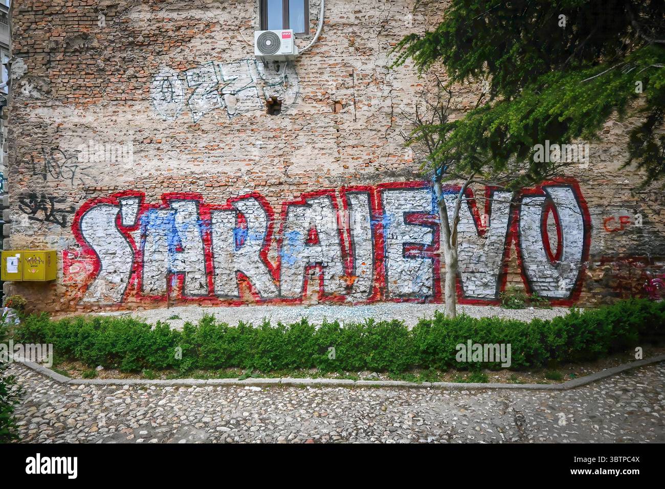 Graffiti-Kunstwerke mit der Schreibweise „Sarajevo“ auf einer alten Ziegelmauer in einem städtischen Gebiet, umgeben von Kopfsteinpflaster und Grün. Stockfoto