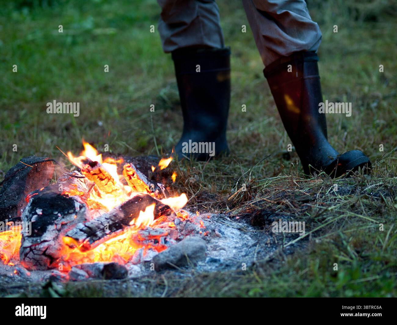 Helles Lagerfeuer, das mit glühenden Baumstämmen und Gluten in einem Wald brennt, während eine Person in der Nähe steht – ein warmer Moment in der Wildnis. Stockfoto Helles Lagerfeuer, das mit glühenden Baumstämmen und Gluten in einem Wald brennt, während eine Person in der Nähe steht – ein warmer Moment in der Wildnis. Stockfoto