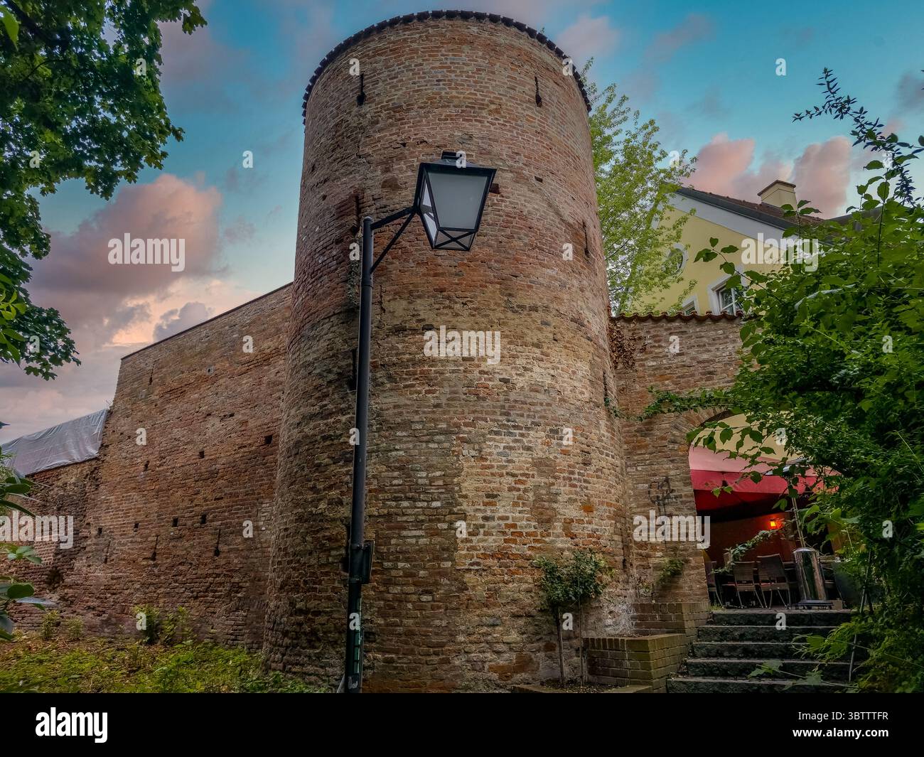 Reste der mittelalterlichen Backsteinmauer und des runden Turms, Schlupflöcher, Sonnenuntergangshimmel mit Wolken in Erding Stockfoto