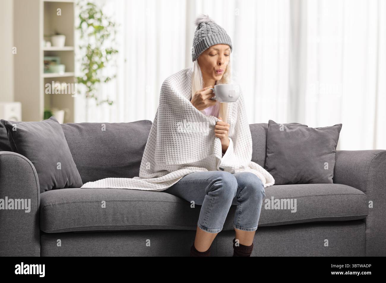 Eine kranke Frau, in eine Decke gewickelt, sitzt auf einem Sofa und hält eine Tasse heißes Getränk Stockfoto