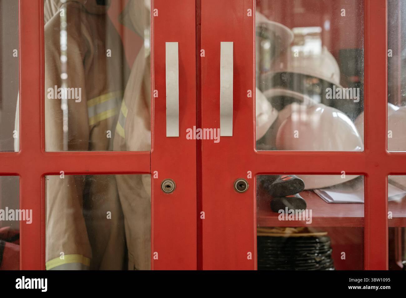 Nahaufnahme eines roten Brandschutzschranks mit Feuerwehruniform, Stiefeln und Helmen hinter Glastüren. Stockfoto
