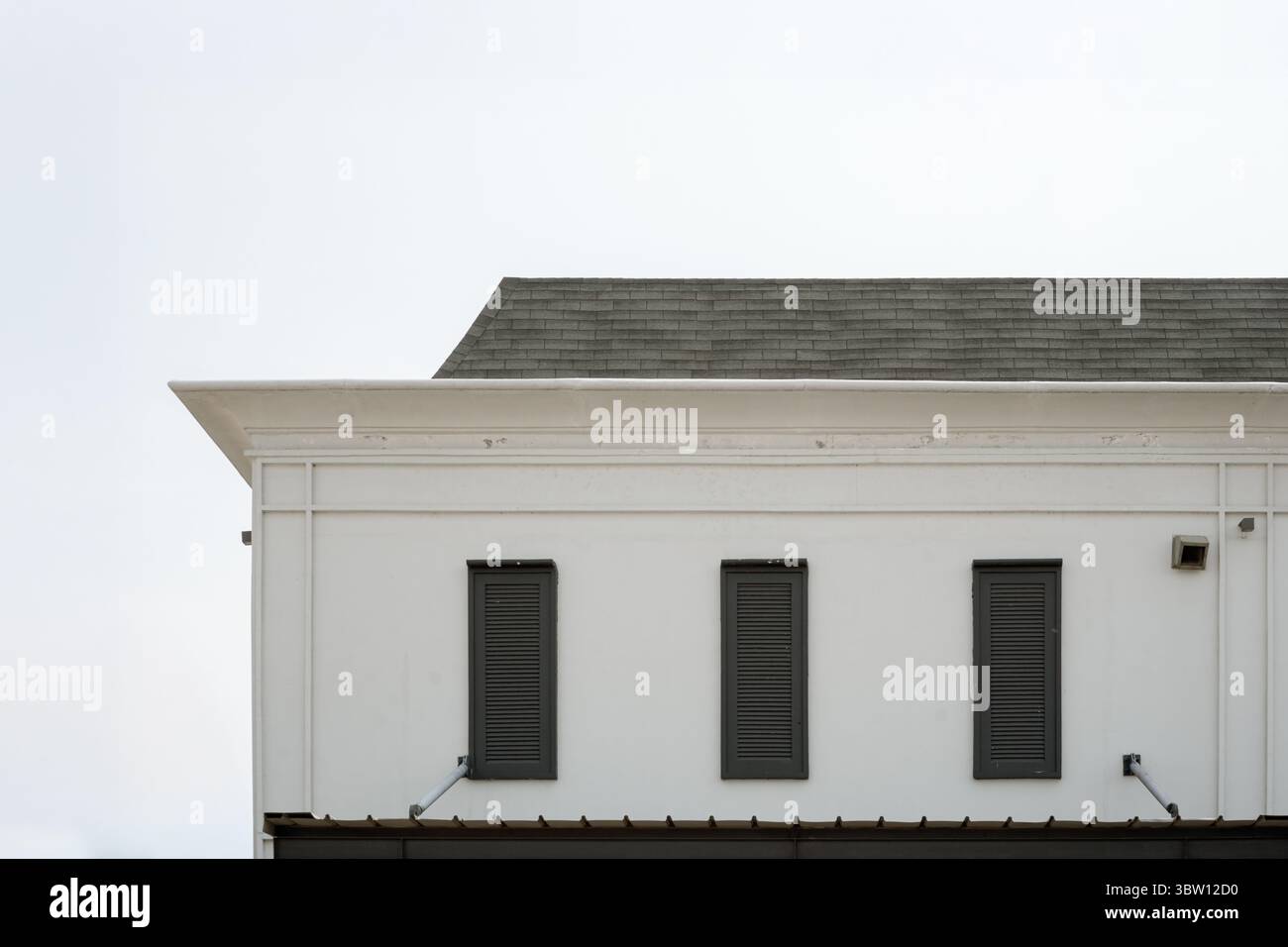 Minimalistisches architektonisches Detail einer weißen Gebäudefassade mit drei dunklen Fenstern und einem grauen Schindeldach vor einem sauberen weißen Himmel. Stockfoto